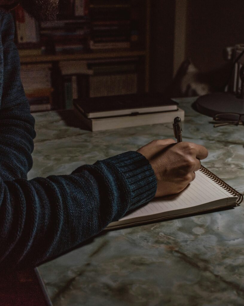 A man sitting alone at a desk in front of a window, writing his thoughts in a journal to practice self-awareness and overcome the fear of solitude.