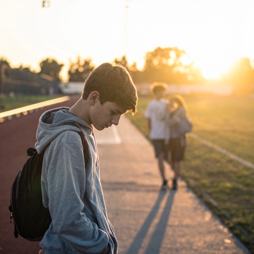 A lonely teenage boy looking down in the foreground while a blurred couple walks together in the sunny background, symbolizing unrequited love and isolation.