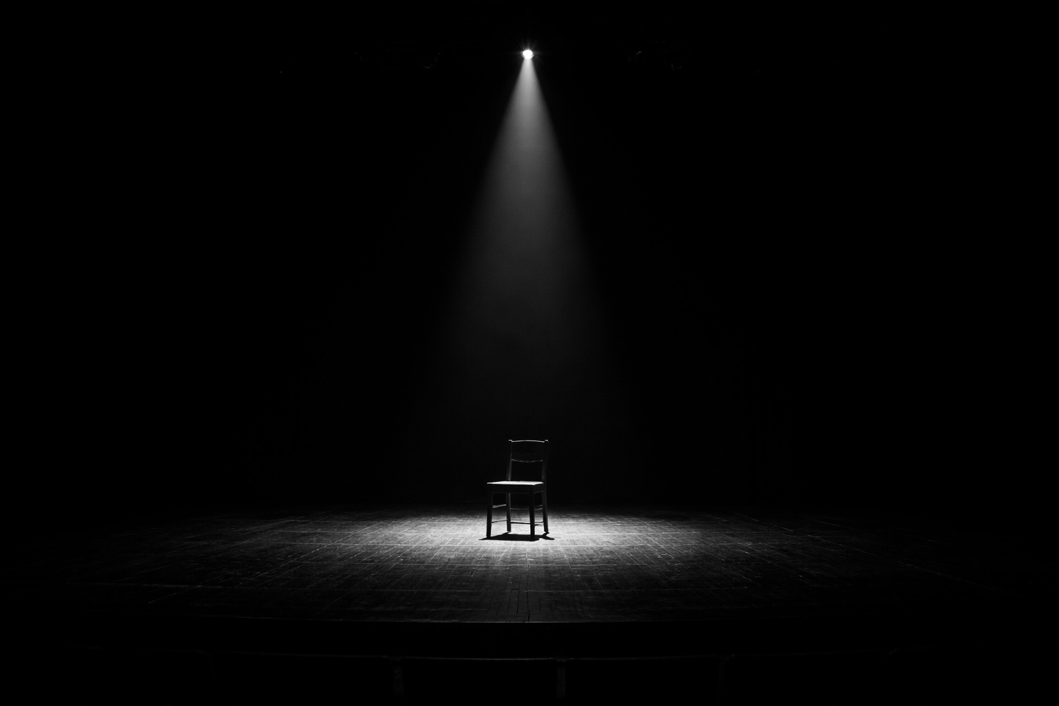 stark, black-and-white photograph of a single empty wooden chair illuminated by a harsh overhead spotlight on a pitch-black stage. This visualizes the Yoga Vasistha concept of Drishti-Srishti, where the mind projects the drama of reality onto an empty void, and the detached action of a Jivanmukta.