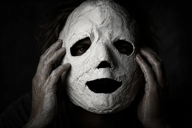 A person with powder-covered hands holding a rough, white plaster mask over their face against a stark black background, symbolizing the facade of societal decency and hidden internal struggles.